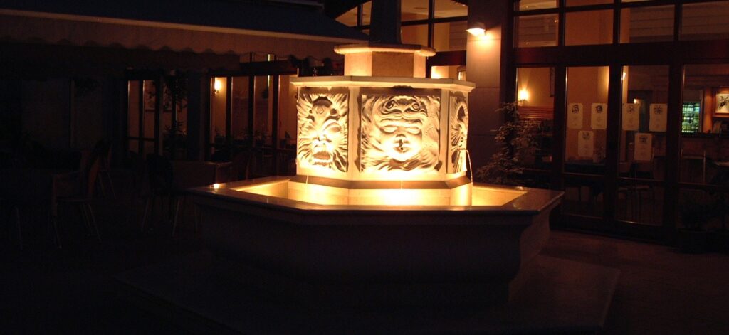 Mészkő szökőkút faragott, arcokat ábrázoló tagozattal éjszaka • A limestone fountain mounted with carved face elements in night light