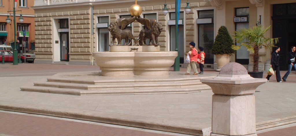 Gyerekek sétálnak a mészkőből faragott szökőkút körül a szegedi Klauzál téren • Childrens walking around the limestone fountain in Klauzal square Szeged -home2